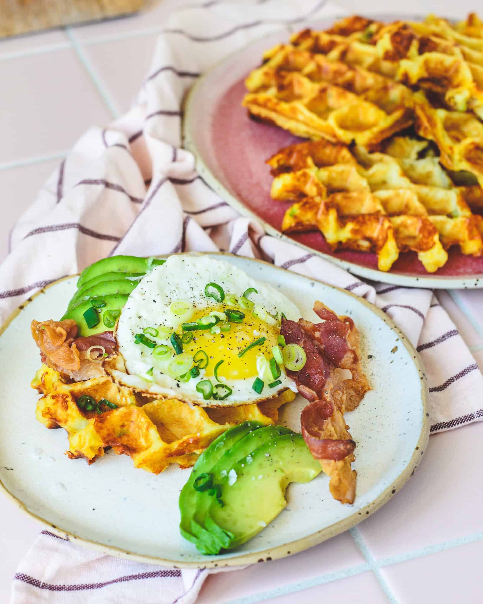 Kartoffelvafler toppet med spejlæg, sprødstegt bacon, frisk avocado og forårsløg serveret på en keramik-tallerken – sprøde og gyldne vafler i baggrunden på en rosa tallerken. Perfekt brunch-ret med cremet og sprød kontrast.
