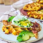 Kartoffelvafler toppet med spejlæg, sprødstegt bacon, frisk avocado og forårsløg serveret på en keramik-tallerken – sprøde og gyldne vafler i baggrunden på en rosa tallerken. Perfekt brunch-ret med cremet og sprød kontrast.