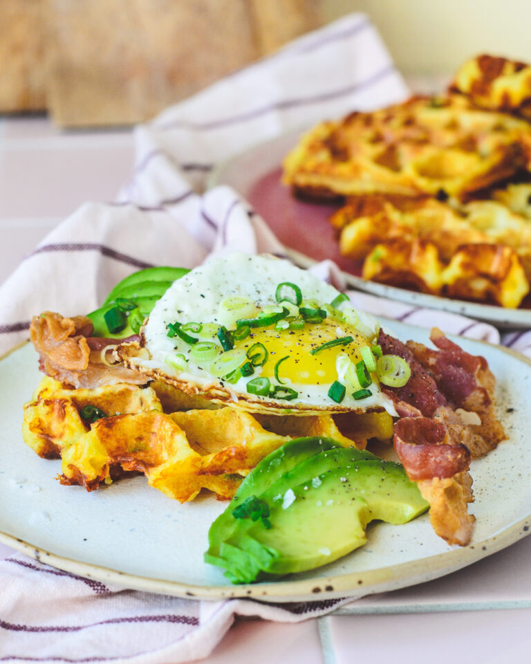 Kartoffelvafler toppet med spejlæg, sprødstegt bacon, frisk avocado og forårsløg serveret på en keramik-tallerken – sprøde og gyldne vafler i baggrunden på en rosa tallerken. Perfekt brunch-ret med cremet og sprød kontrast.