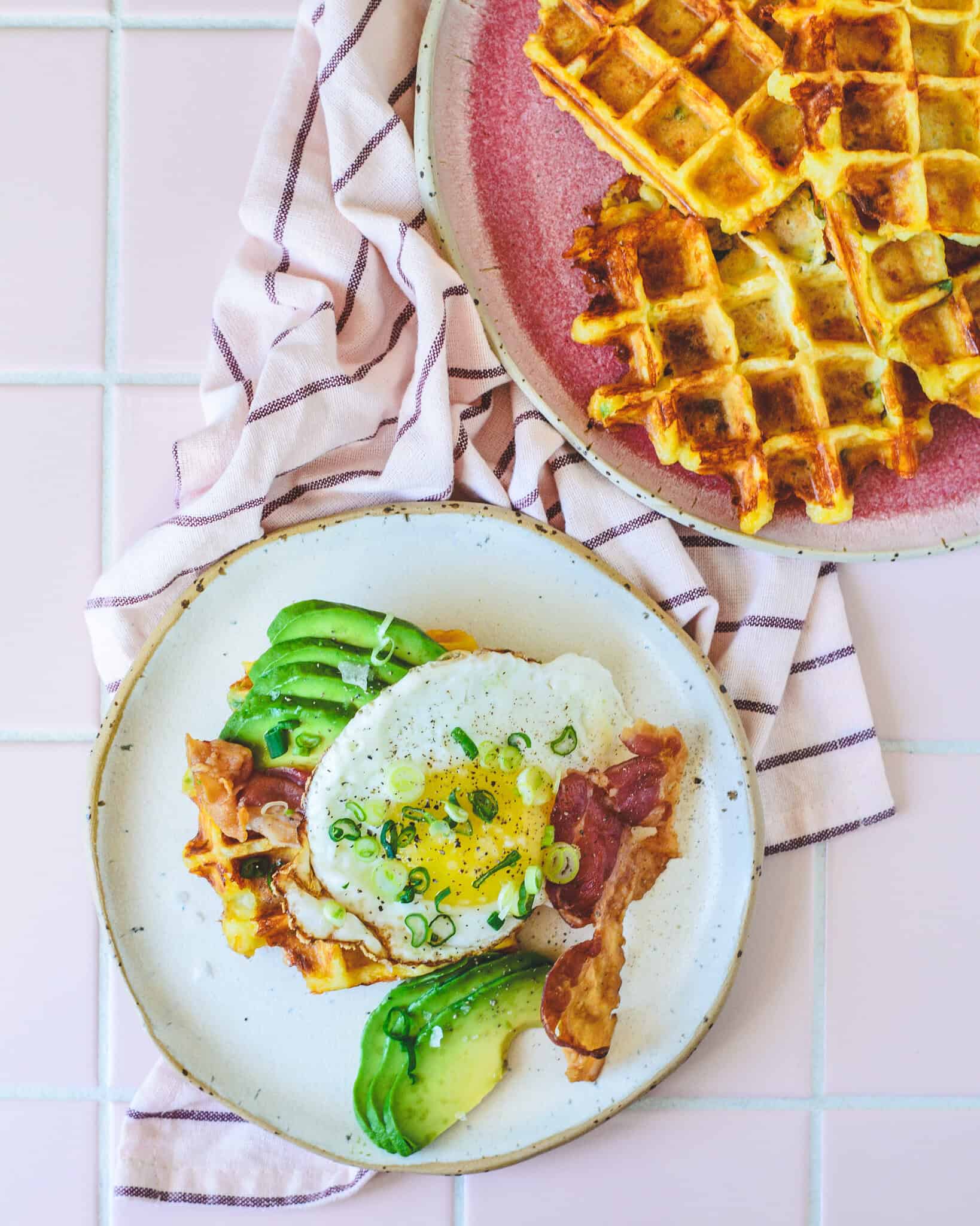 Kartoffelvafler toppet med spejlæg, sprødstegt bacon, frisk avocado og forårsløg serveret på en keramik-tallerken – sprøde og gyldne vafler i baggrunden på en rosa tallerken. Perfekt brunch-ret med cremet og sprød kontrast.