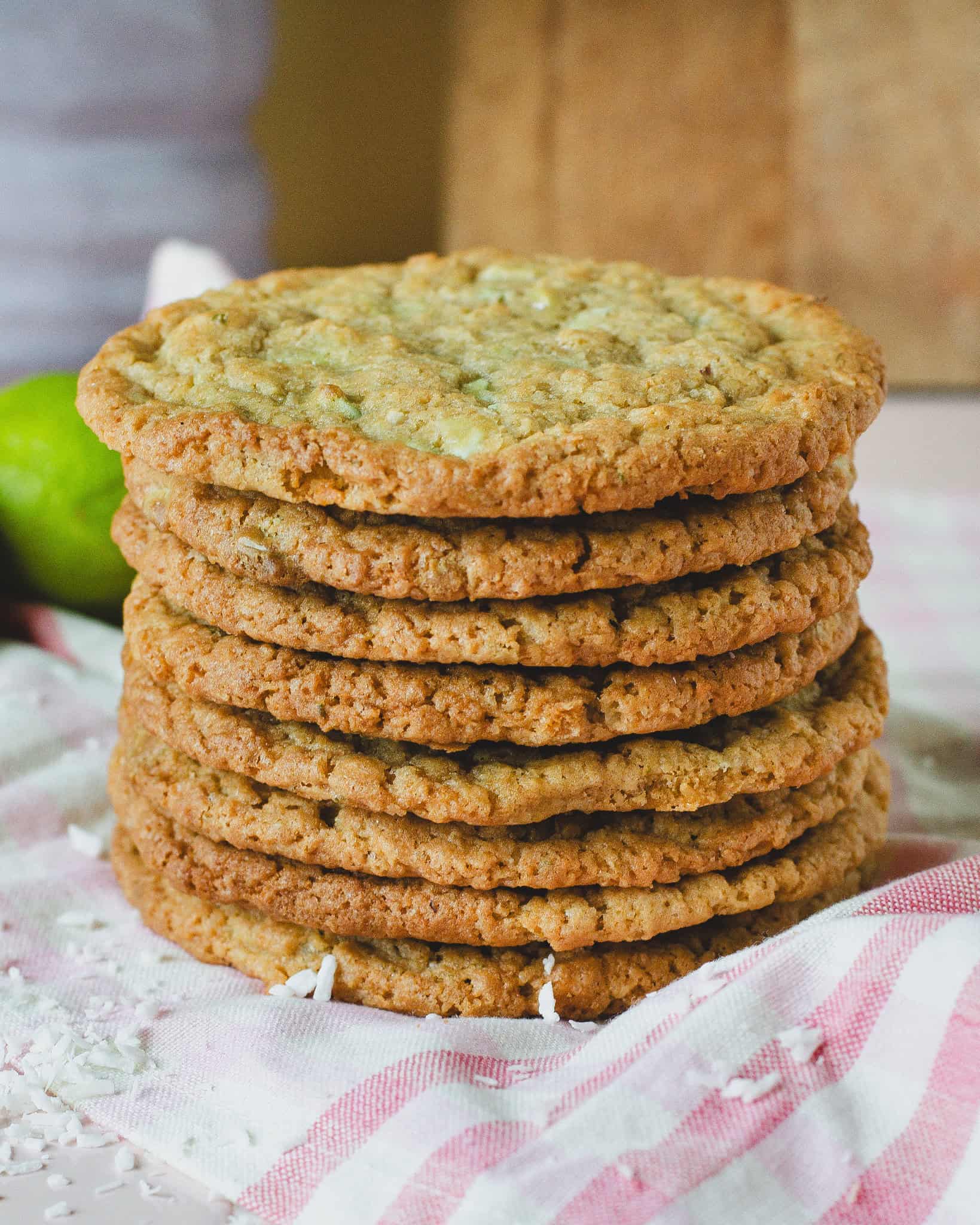 Stabel af sprøde limecookies med kokos og chokolade, serveret på et lyserødt stribet viskestykke. Cookiesene er gyldne og jævnt bagte med tydelige stykker kokos og chokolade, og en frisk lime anes i baggrunden. Perfekt til en sommerkage eller som lækker snack med et tropisk twist.
