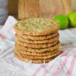 Stabel af sprøde kokos lime cookies, serveret på et lyserødt stribet viskestykke. Cookiesene er gyldne og jævnt bagte med tydelige stykker kokos og chokolade, og en frisk lime anes i baggrunden. Perfekt til en sommerkage eller som lækker snack med et tropisk twist.