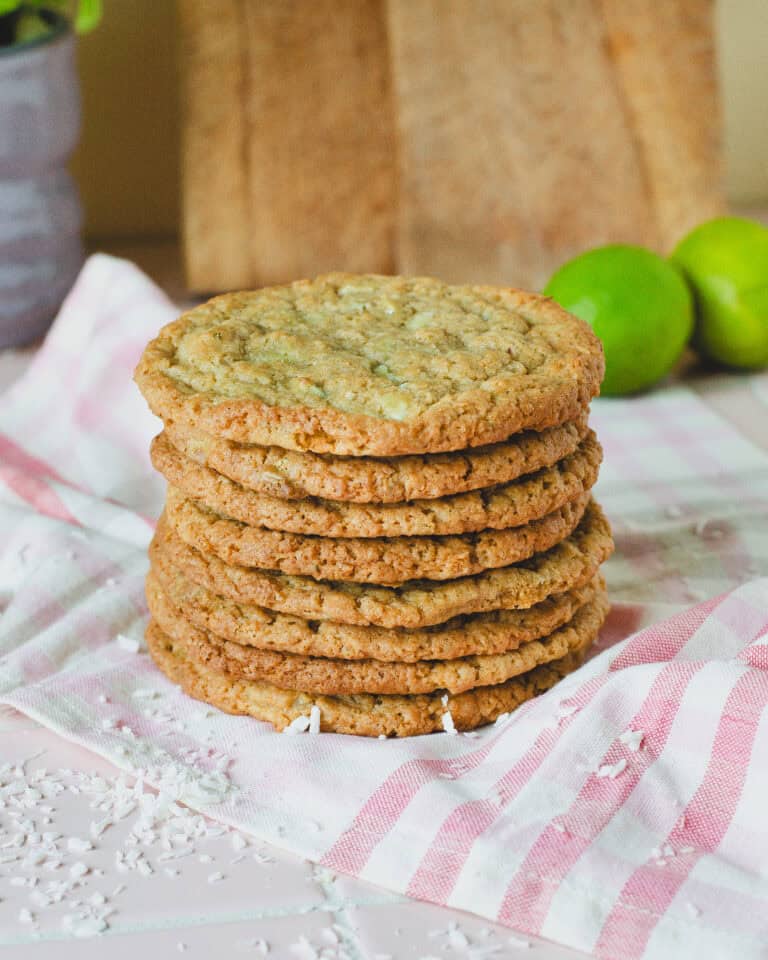 Stabel af sprøde kokos lime cookies, serveret på et lyserødt stribet viskestykke. Cookiesene er gyldne og jævnt bagte med tydelige stykker kokos og chokolade, og en frisk lime anes i baggrunden. Perfekt til en sommerkage eller som lækker snack med et tropisk twist.