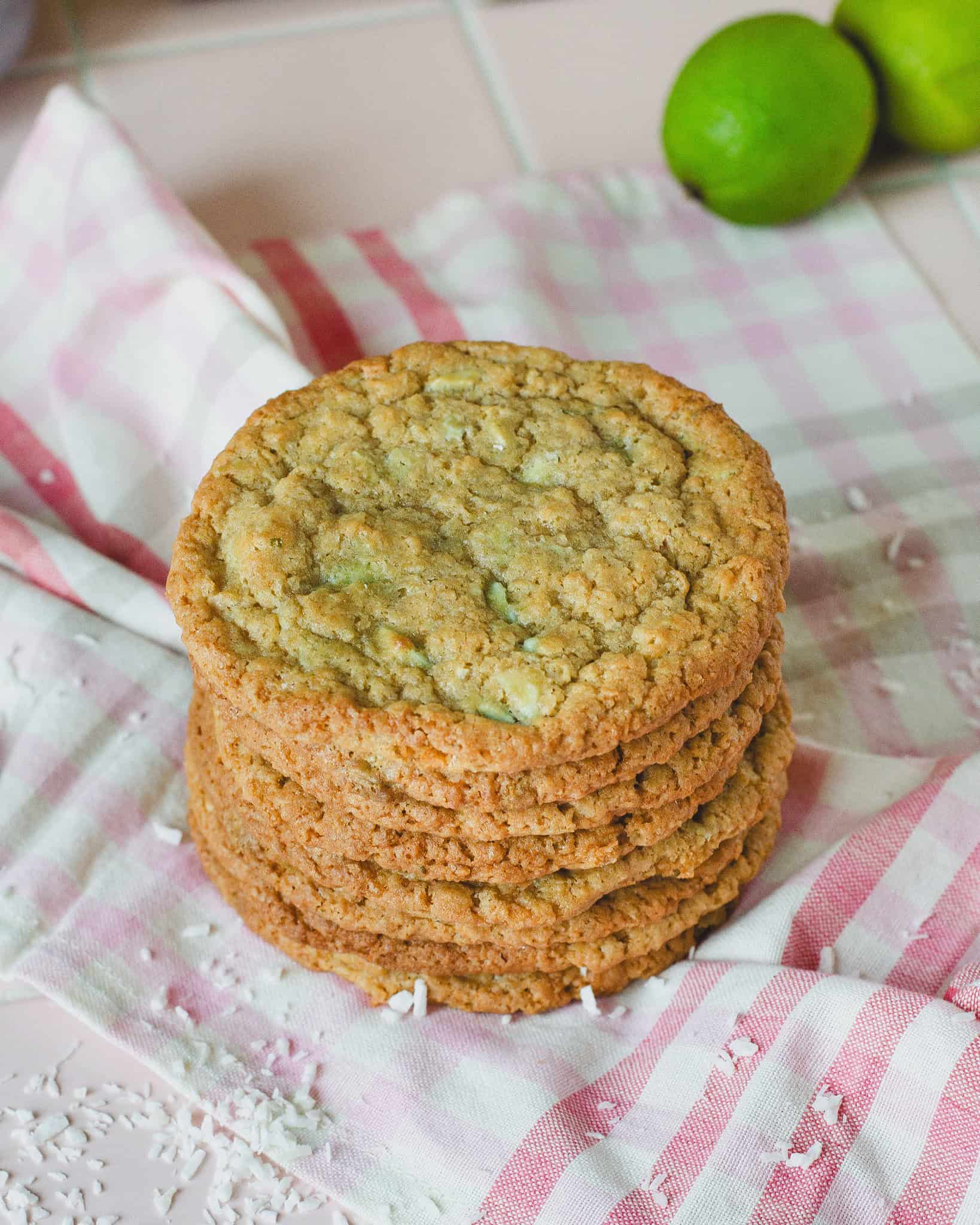 Stabel af sprøde kokos lime cookies, serveret på et lyserødt stribet viskestykke. Cookiesene er gyldne og jævnt bagte med tydelige stykker kokos og chokolade, og en frisk lime anes i baggrunden. Perfekt til en sommerkage eller som lækker snack med et tropisk twist.