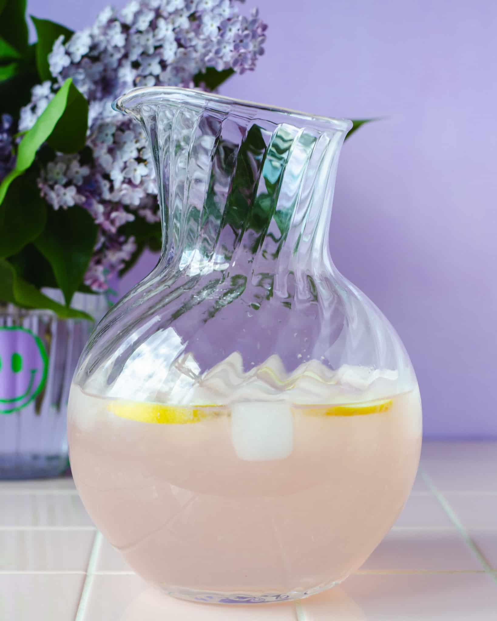 En glaskande syren lemonade  med isterninger, citronskive placeret foran en vase med friske syrener. Stemningsfuld og sommerlig scene i lilla nuancer.