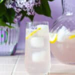 Et højt glas syrenlimonade med isterninger, citronskive og en syrenblomst i toppen, placeret foran en glaskande med mere limonade. I baggrunden står en vase med friske syrener. Stemningsfuld og sommerlig scene i lilla nuancer.