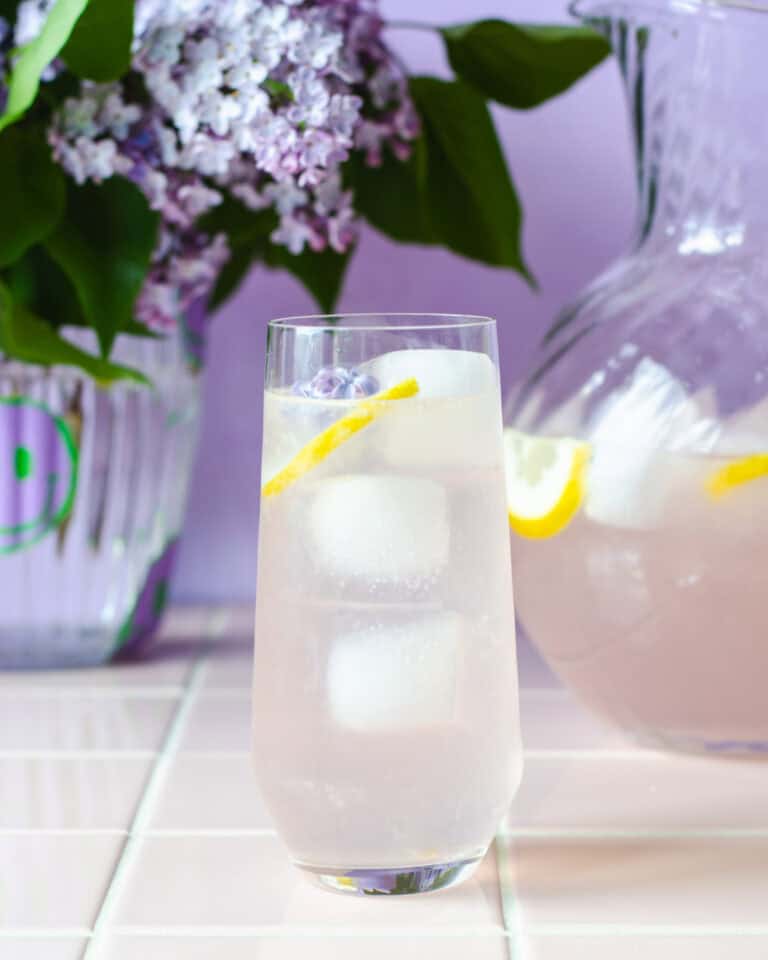 Et højt glas syrenlimonade med isterninger, citronskive og en syrenblomst i toppen, placeret foran en glaskande med mere limonade. I baggrunden står en vase med friske syrener. Stemningsfuld og sommerlig scene i lilla nuancer.