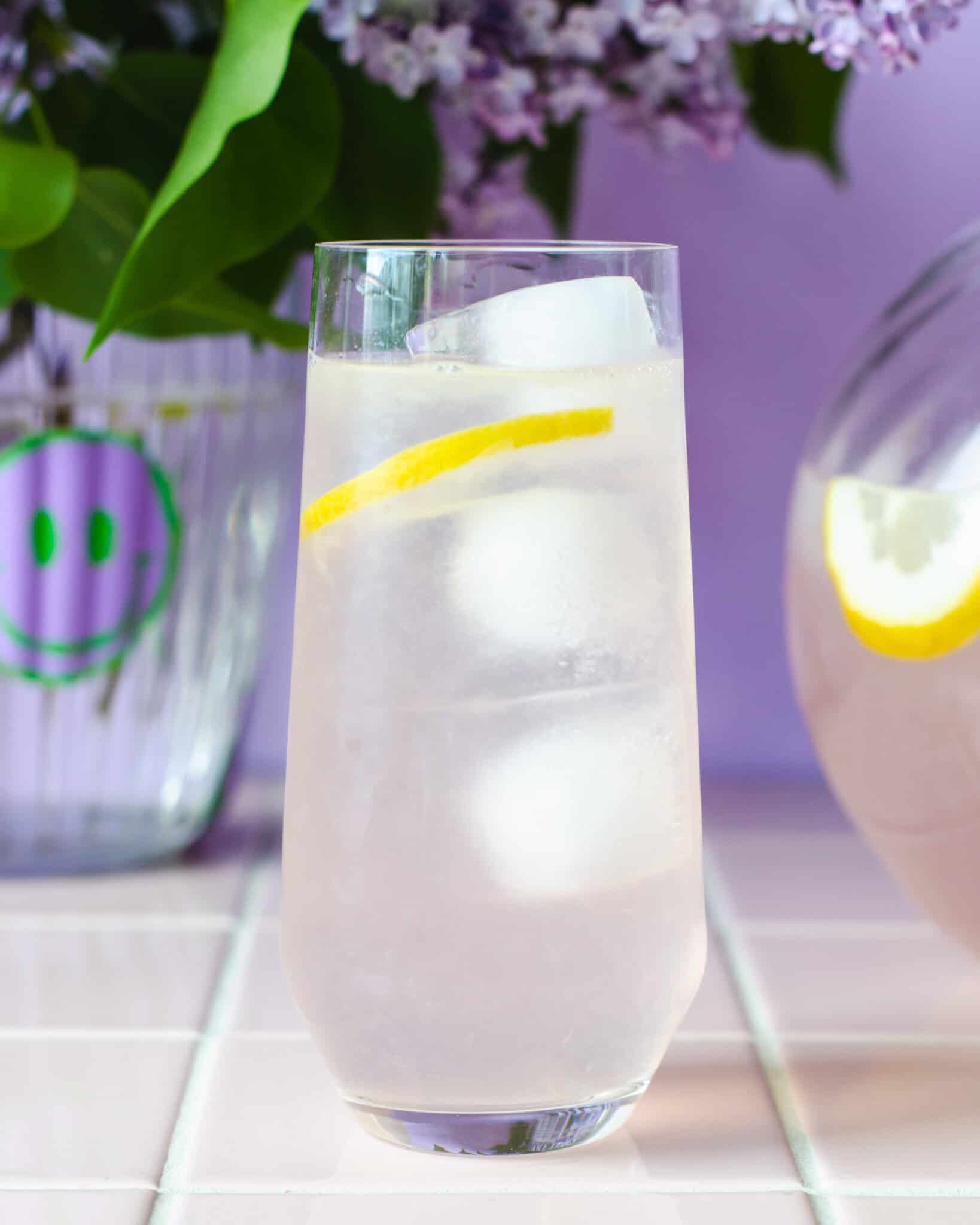 Et højt glas syren lemonade  med isterninger, citronskive og en syrenblomst i toppen, placeret foran en glaskande med mere limonade. I baggrunden står en vase med friske syrener. Stemningsfuld og sommerlig scene i lilla nuancer.