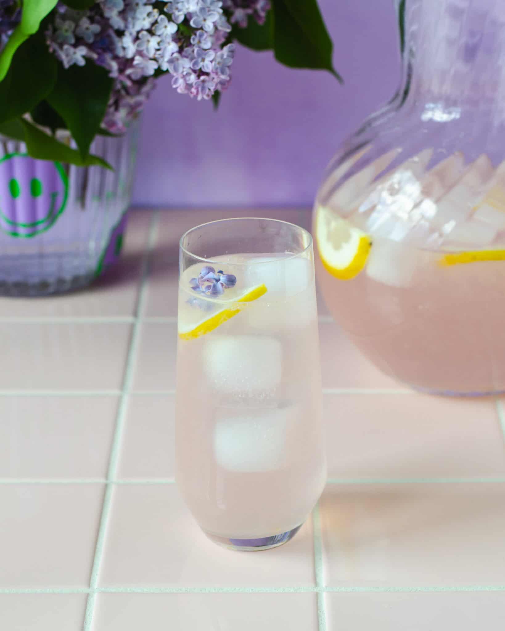 Et højt glas syren lemonade med isterninger, citronskive og en syrenblomst i toppen, placeret foran en glaskande med mere limonade. I baggrunden står en vase med friske syrener. Stemningsfuld og sommerlig scene i lilla nuancer.