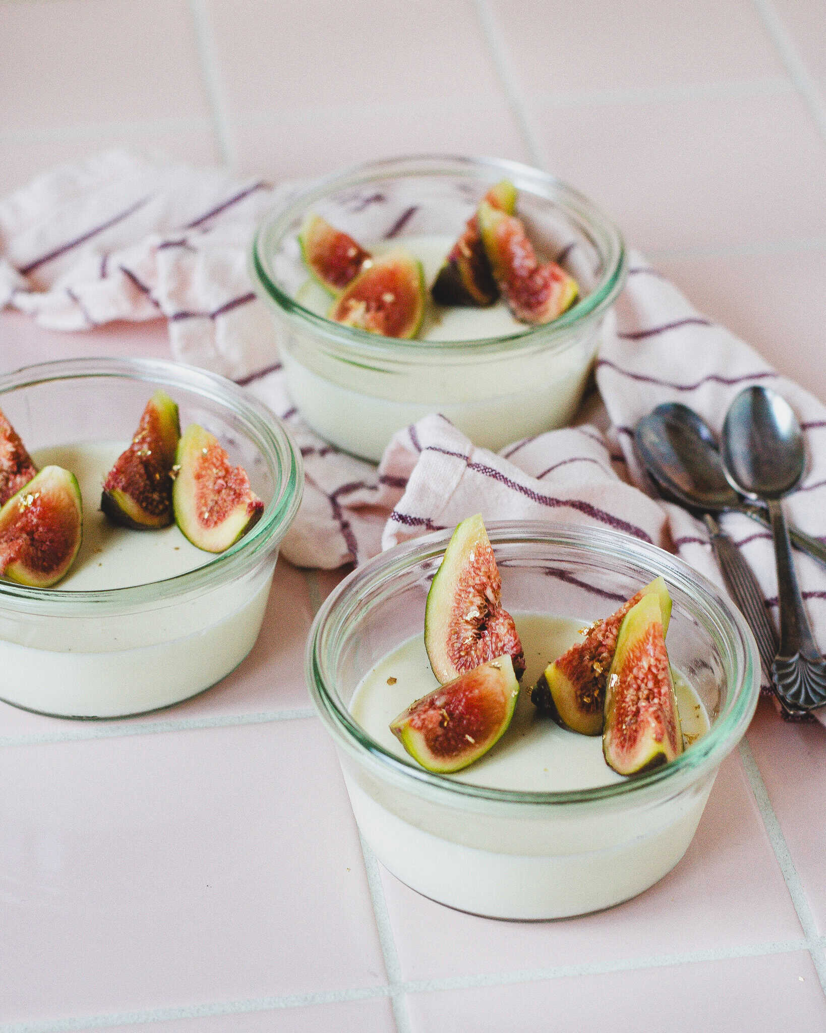 Cremet panna cotta med figenblade toppet med friske figner, serveret i små glas på lyserød bordplade.