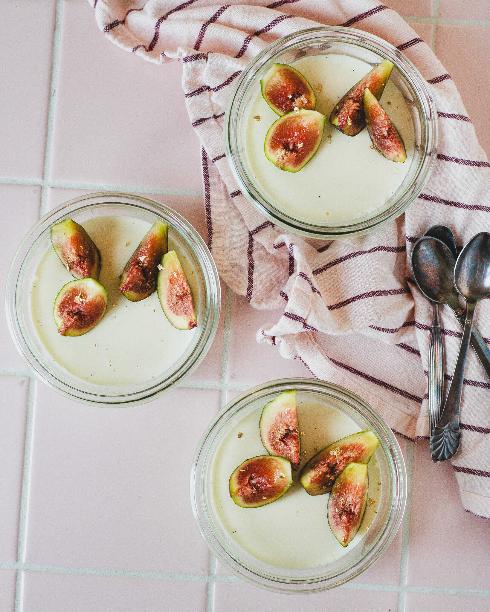 Tre portioner panna cotta med figenblade pyntet med figner, fotograferet på lyserødt bord med viskestykke og skeer.