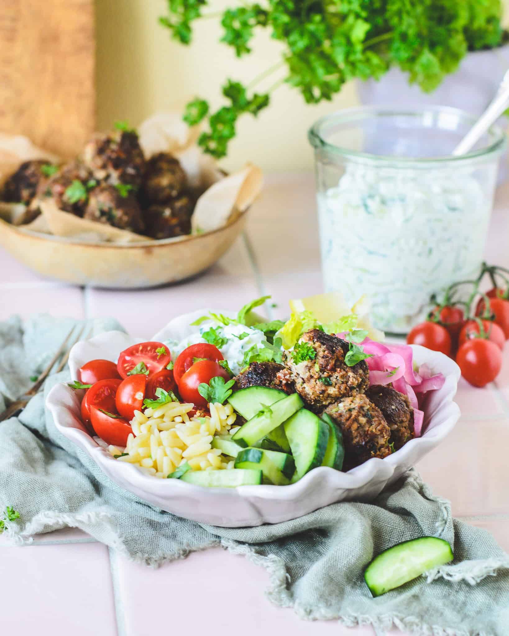 Græsk bowl med kødboller, tzatziki, orzo, agurk, cherrytomater og syltede rødløg serveret i en hvid skål.