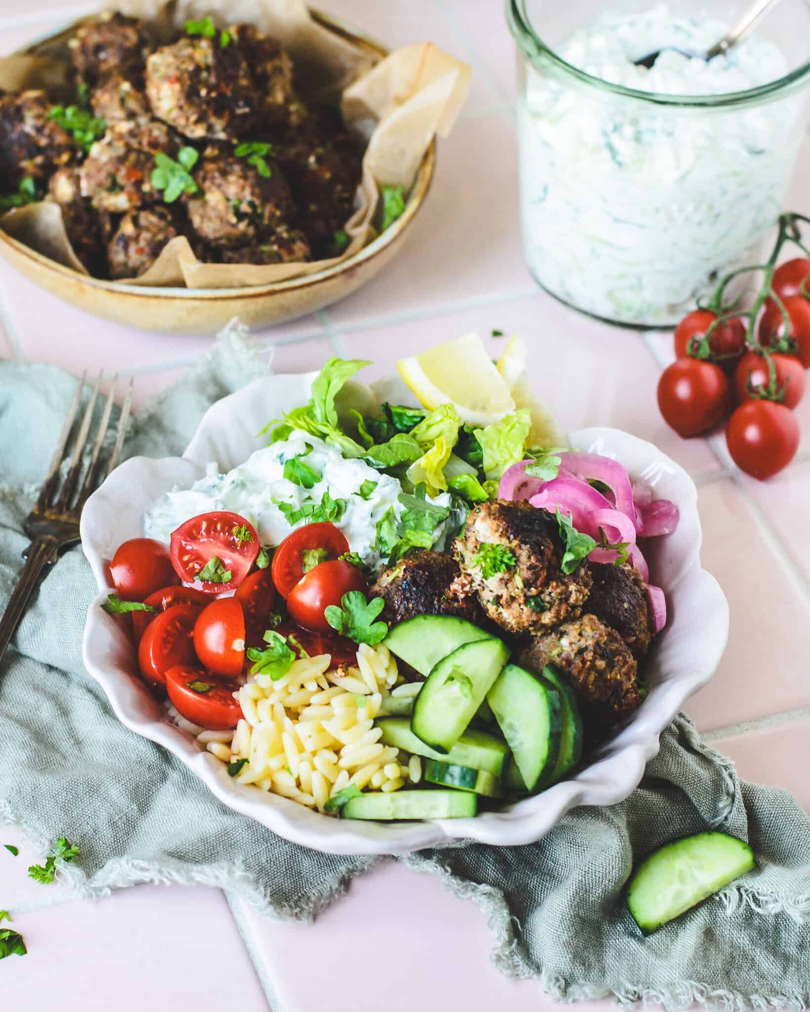 Græsk bowl med hjemmelavede kødboller, cremet tzatziki og orzo serveret med friske grøntsager og citron.