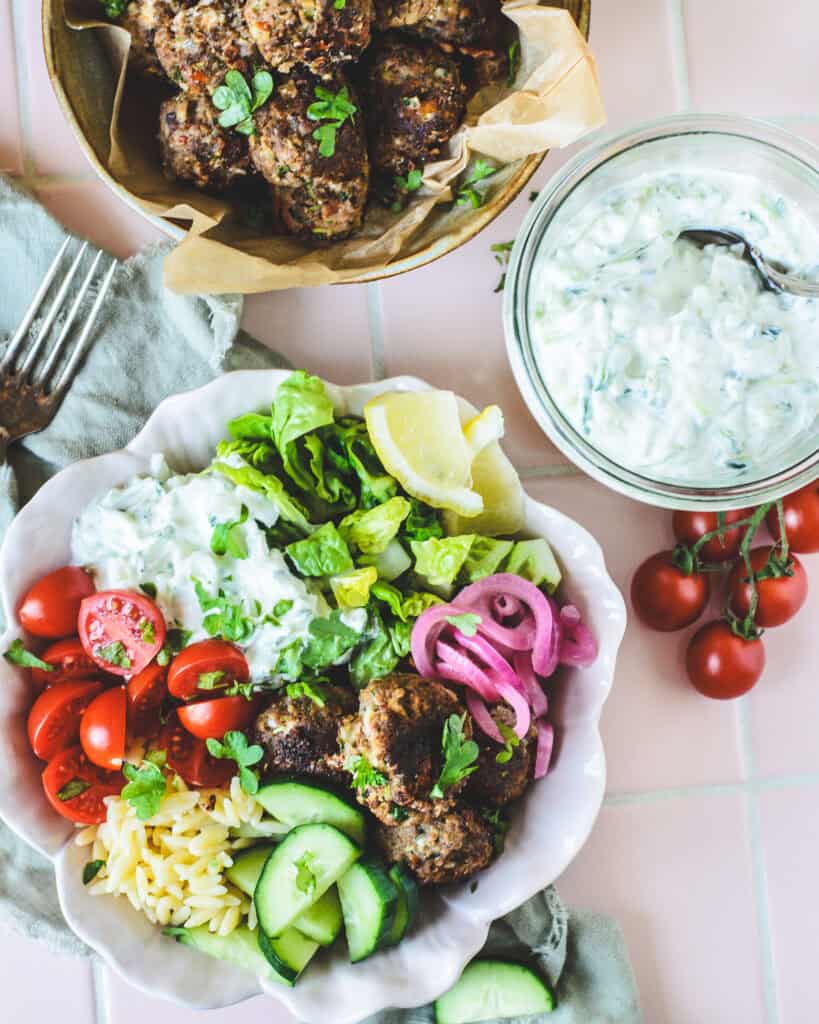 Græsk bowl med kødboller, tzatziki og orzo fotograferet ovenfra med ekstra kødboller og tzatziki i baggrunden.